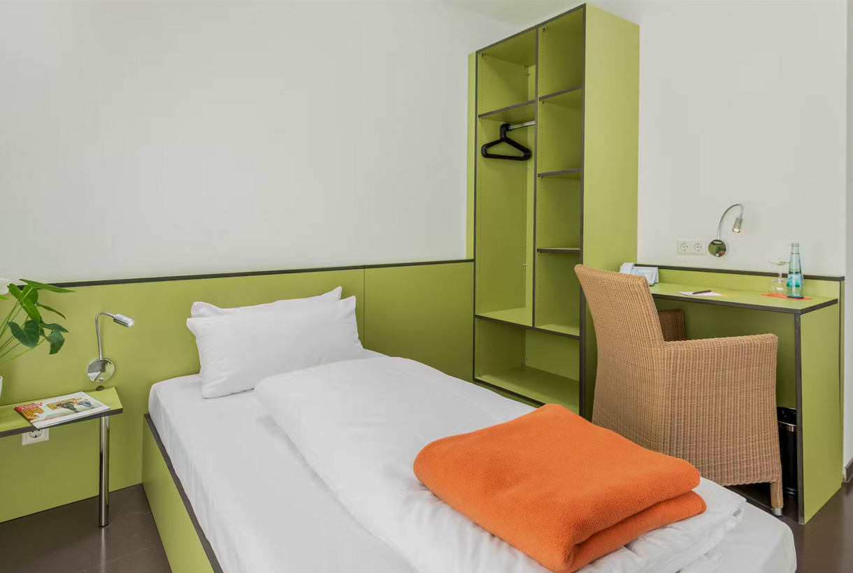 Modernes Einzelzimmer mit Bett, Schreibtisch, Korbsessel und grünem Schrank